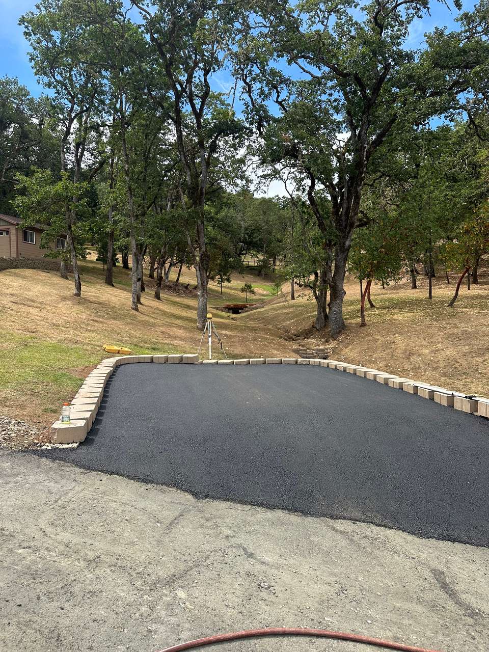 New asphalt pad with a clean edge against a block border, oak trees beyond