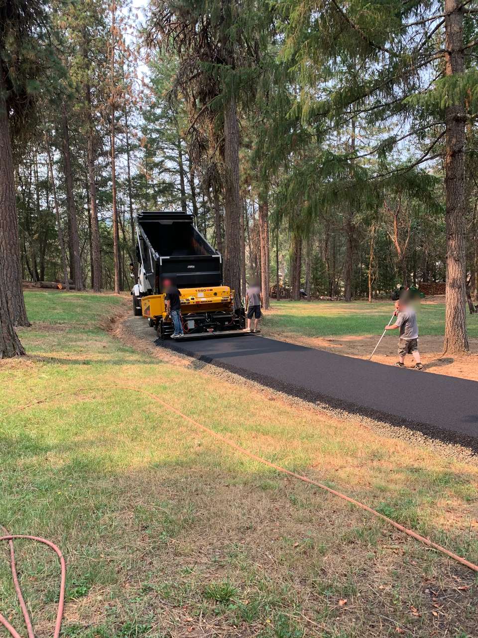 Paving machine laying a new asphalt driveway through a grassy, wooded lot