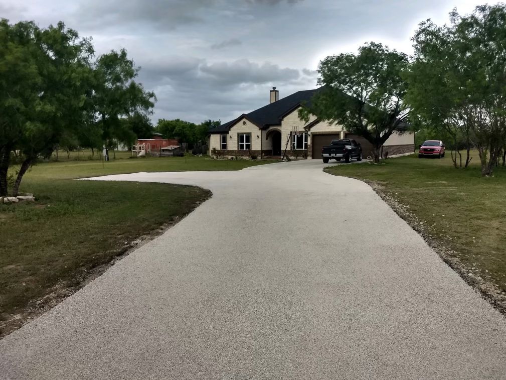 Wide chip seal driveway curving toward a brick home under a cloudy sky, with grass and trees on each side